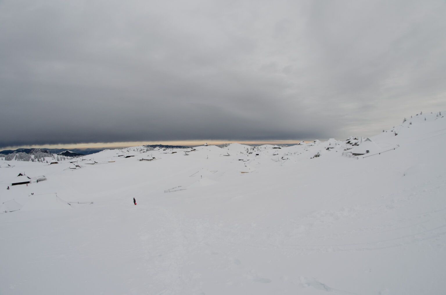 Na zdjęciu widać człowieka stojącego na śniegu, w tle szałasy pasterskie pokryte śniegiem na Wielkiej Planinie